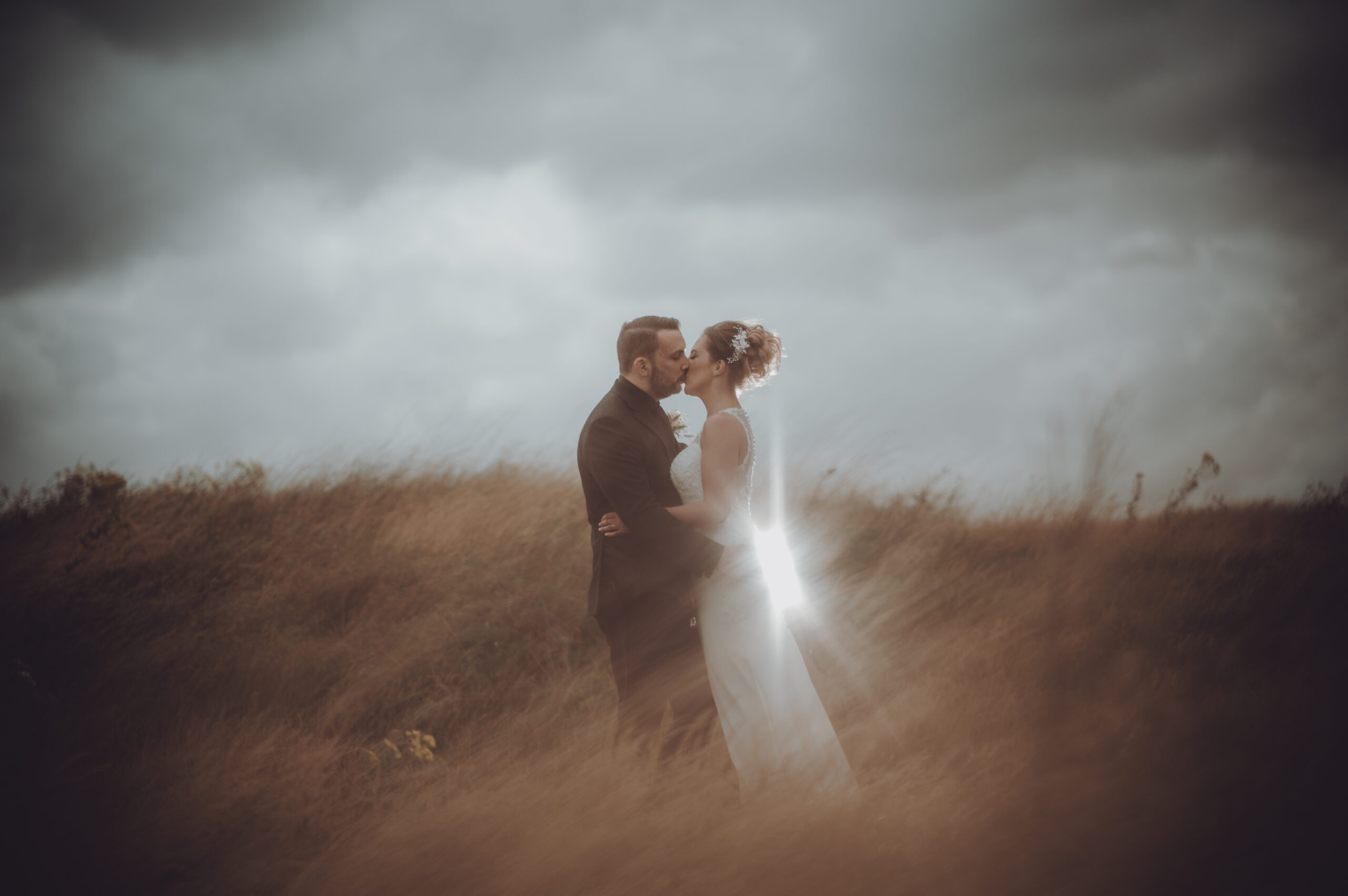 A bride and groom kissing in a field under a stormy sky. (listing)