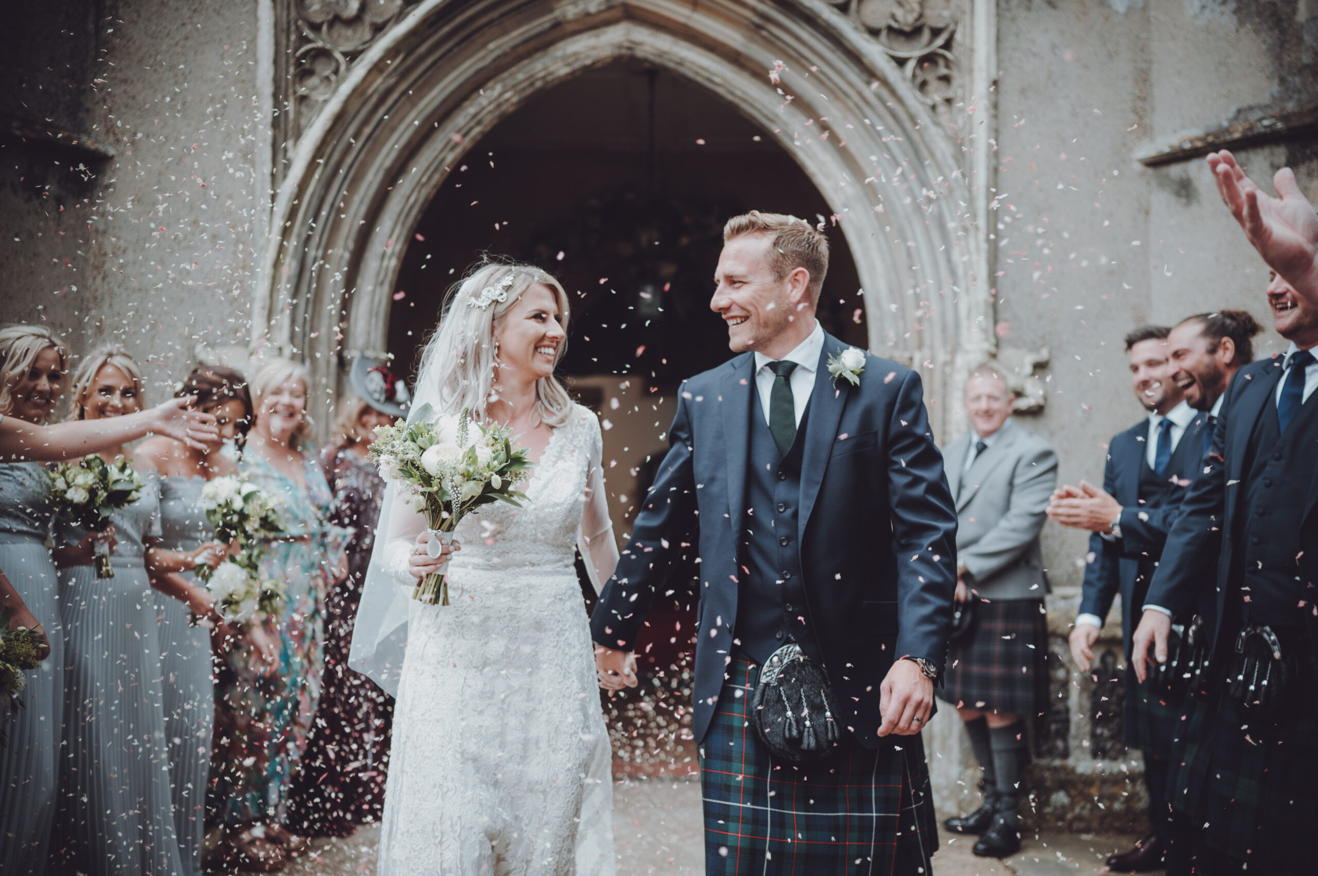 A bride and groom walk out of a church with confetti thrown at them. (listing)