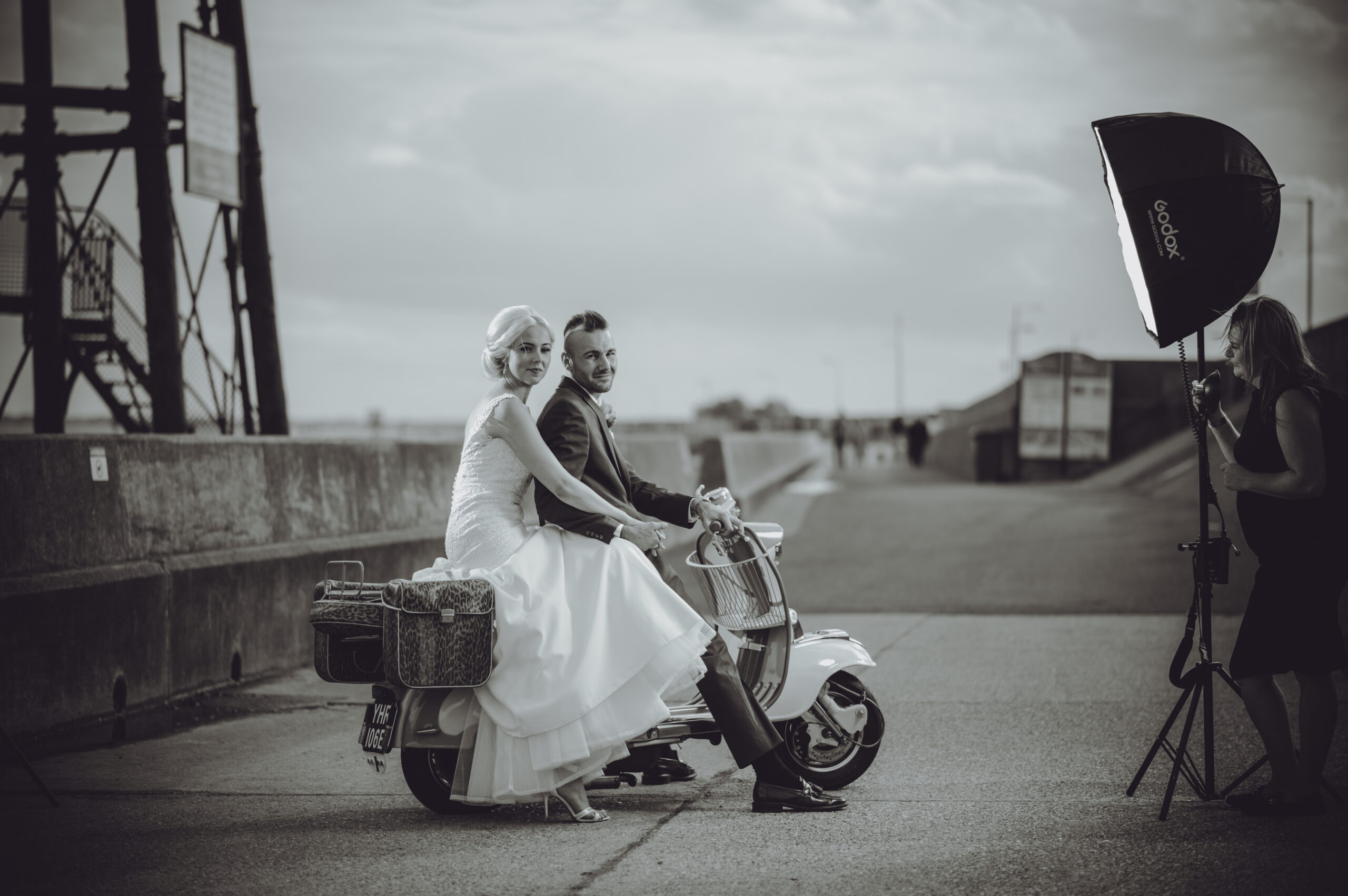 A bride and groom on a scooter in front of a bridge. (listing)