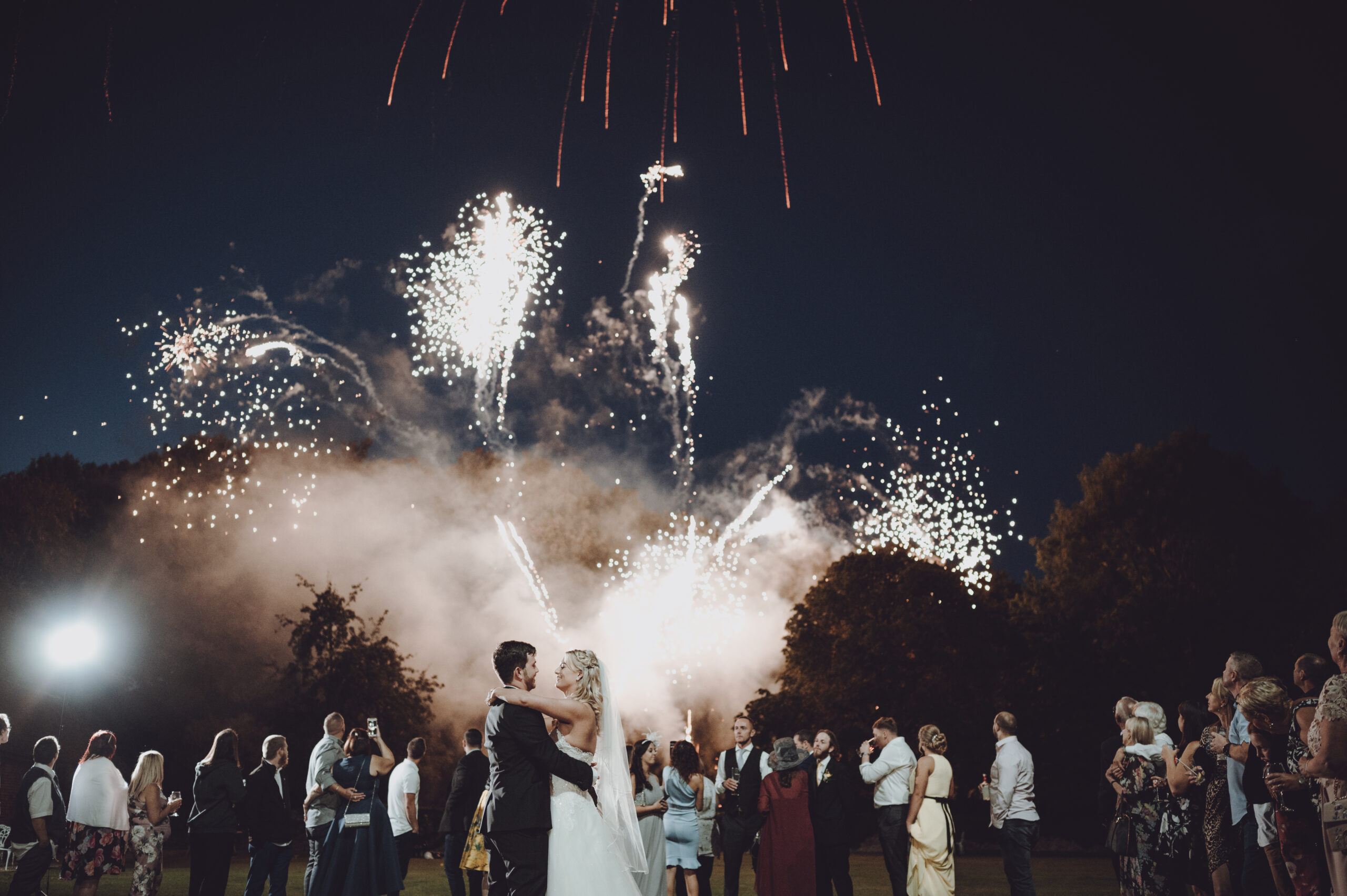 A bride and groom kissing in front of a firework display. (listing)