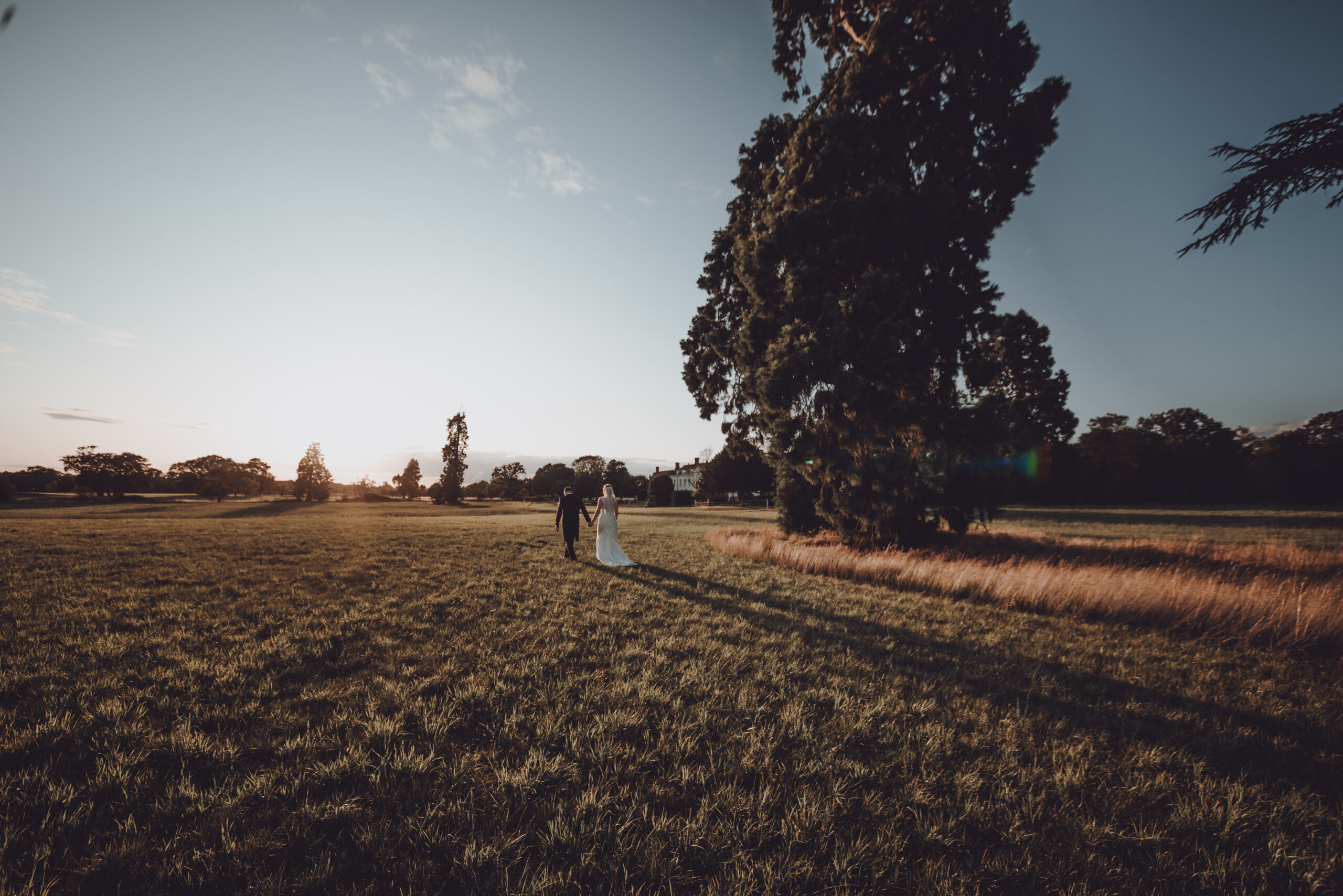 A bride and groom walking in a field at sunset. (listing)