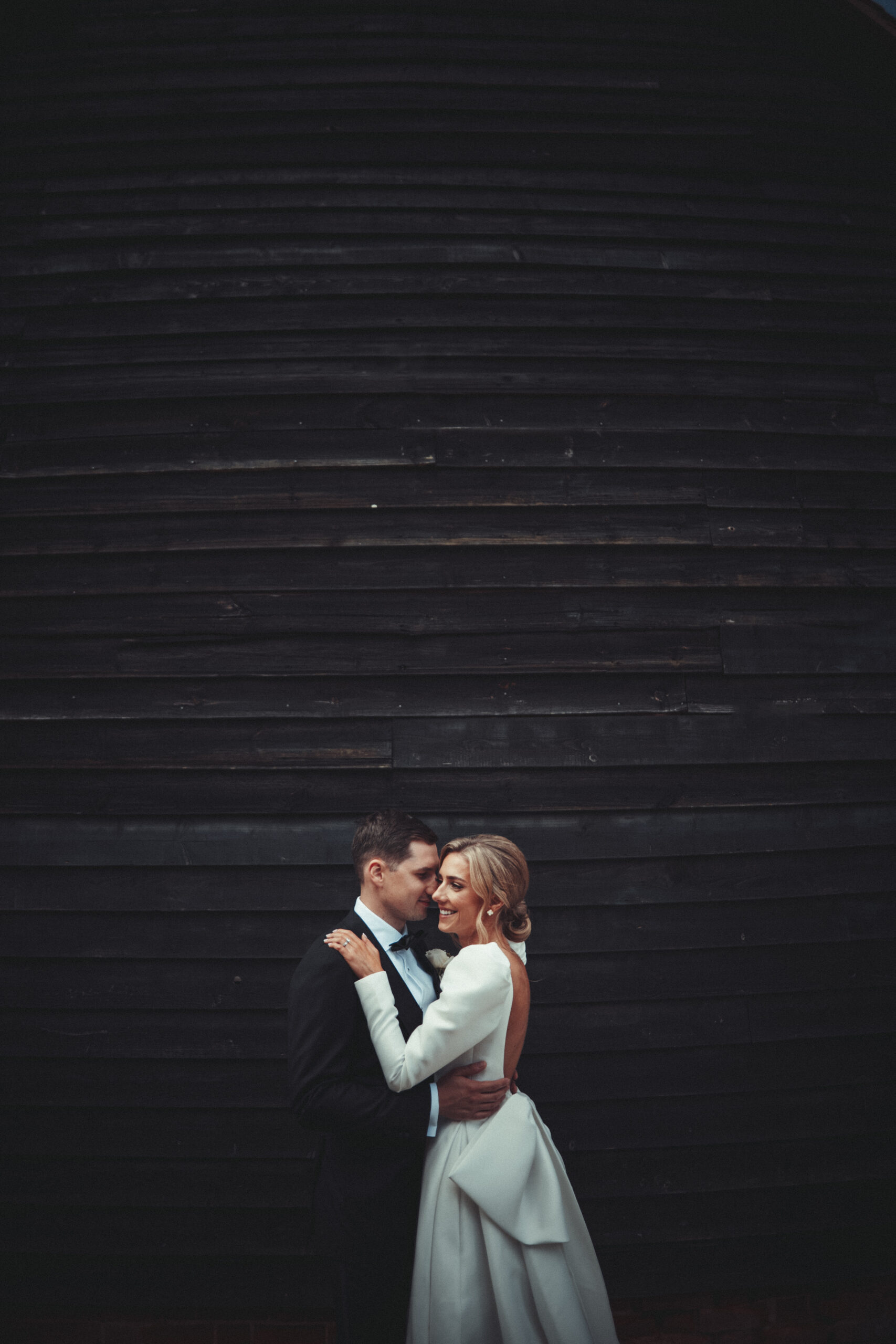 A bride and groom embracing in front of a black barn. (listing)