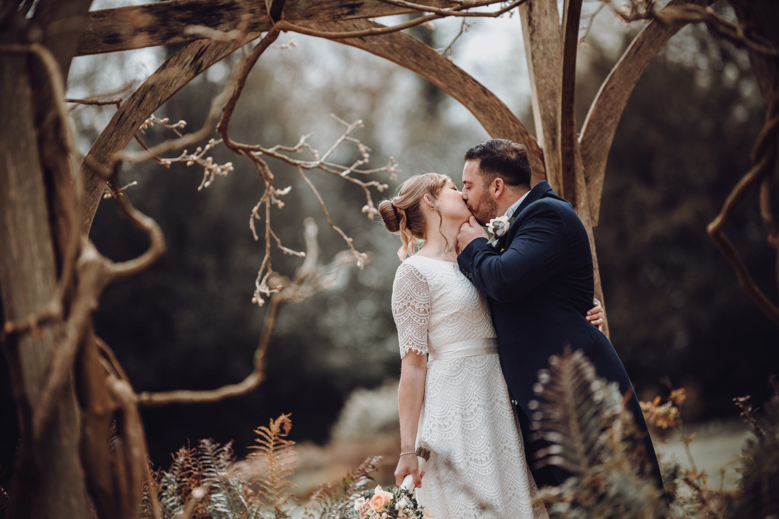 A bride and groom kissing under an arbour in the woods. (listing)