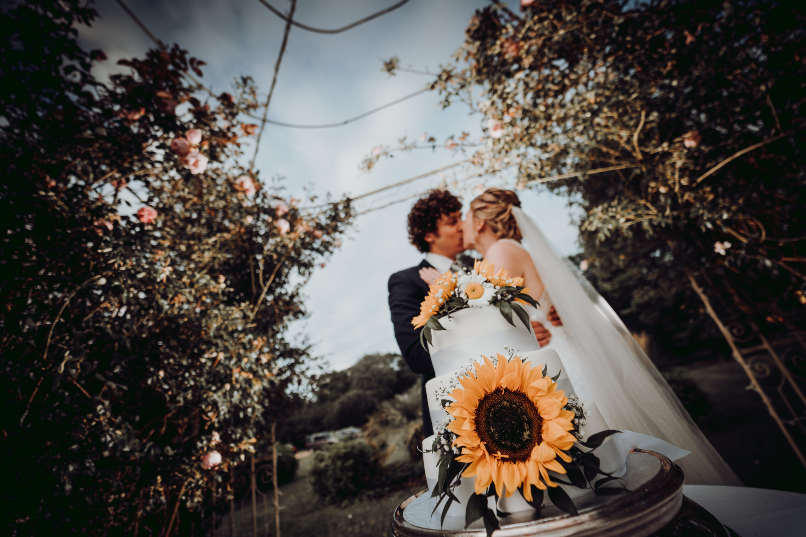 A bride and groom standing next to a wedding cake with sunflowers. (listing)