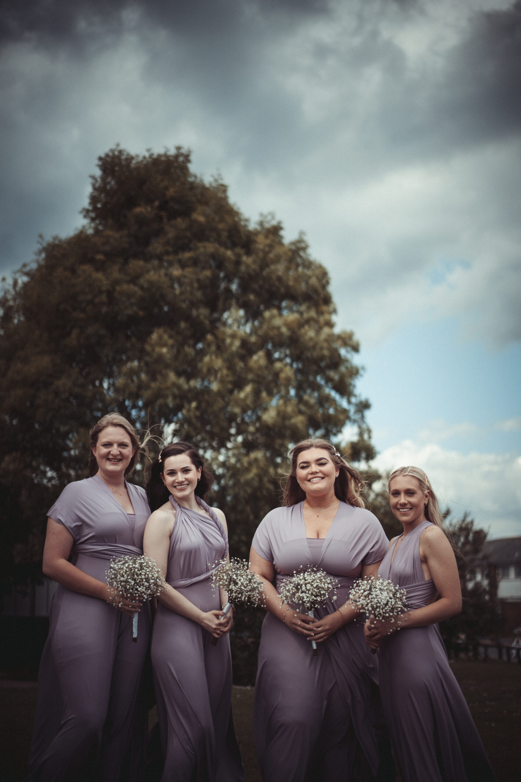 Bridesmaids in lavender dresses standing in front of a cloudy sky. (listing)