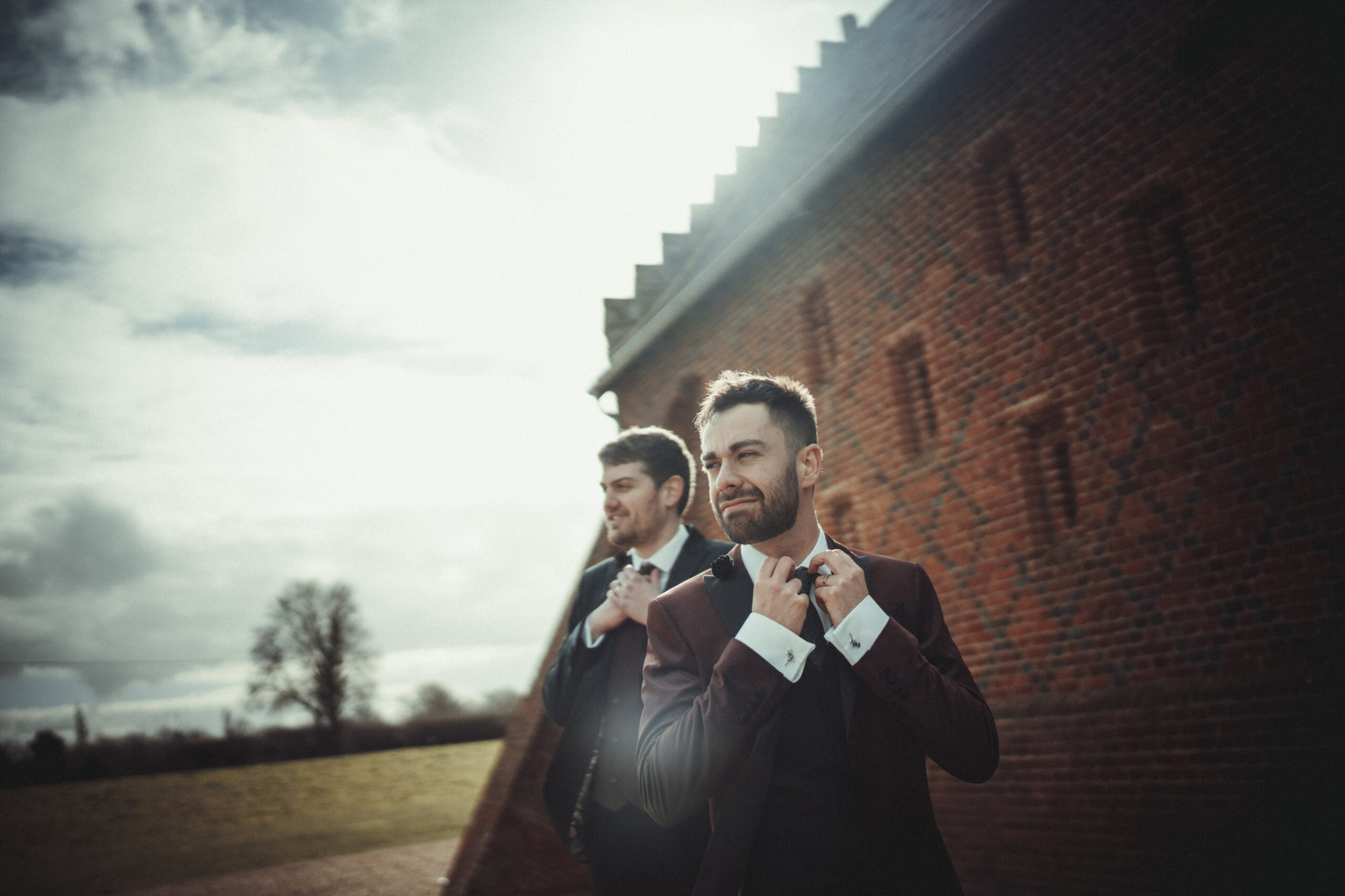 Two men tying their ties in front of a brick wall. (listing)