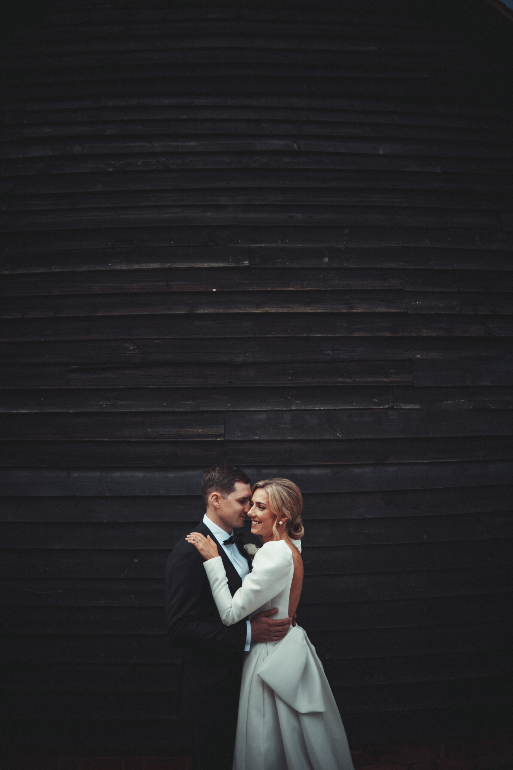 A bride and groom embracing in front of a black barn. (listing)