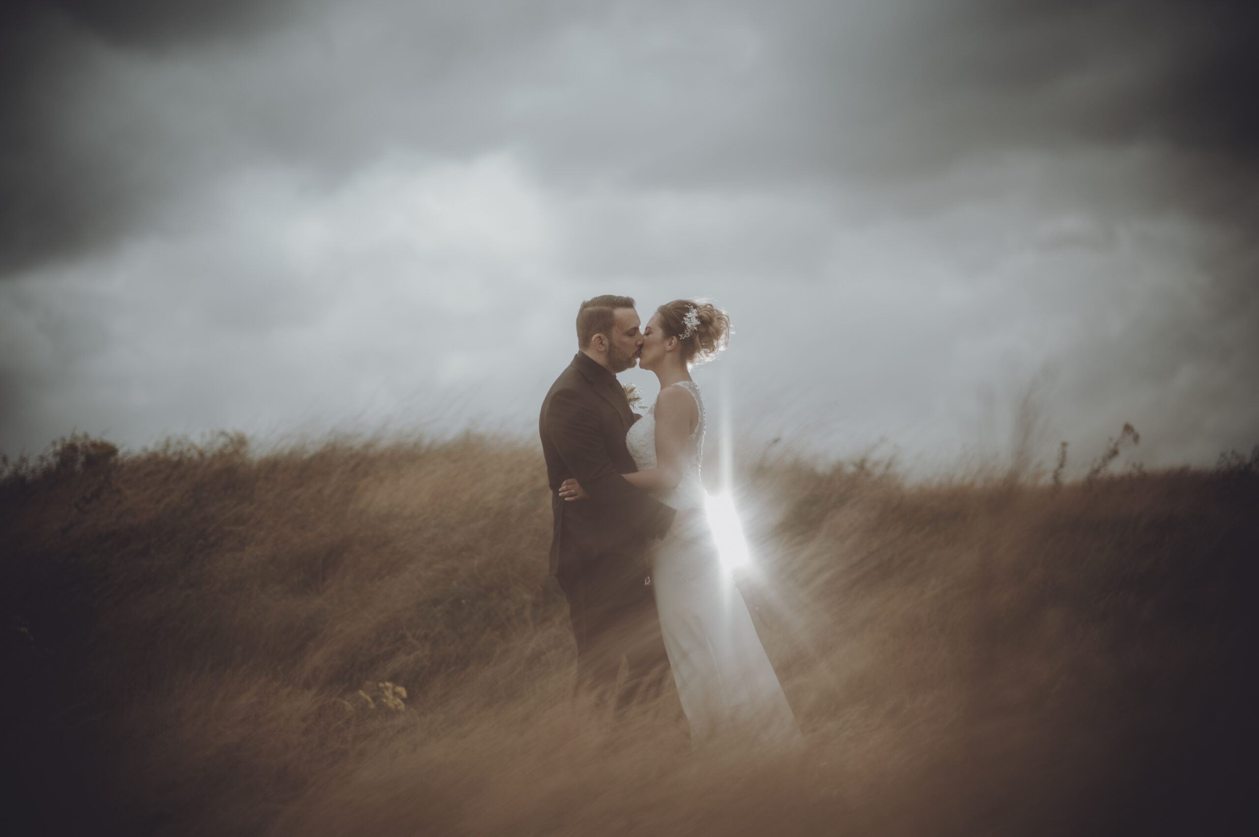 A bride and groom kissing in a field under a stormy sky. (listing)