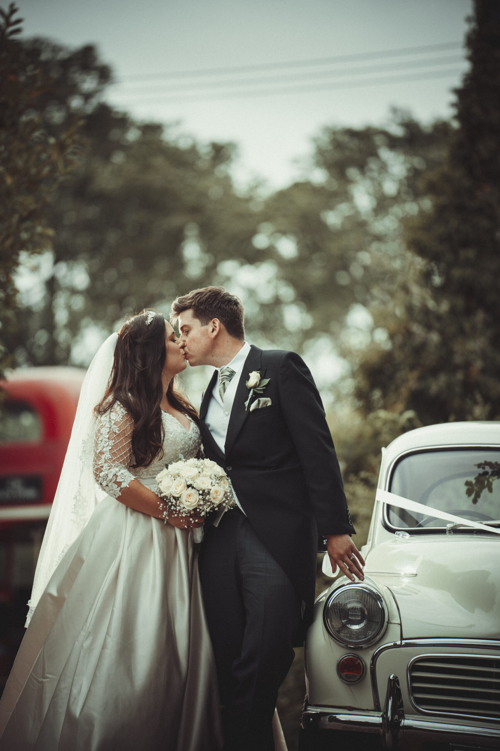 A bride and groom kiss in front of a classic car. (listing)