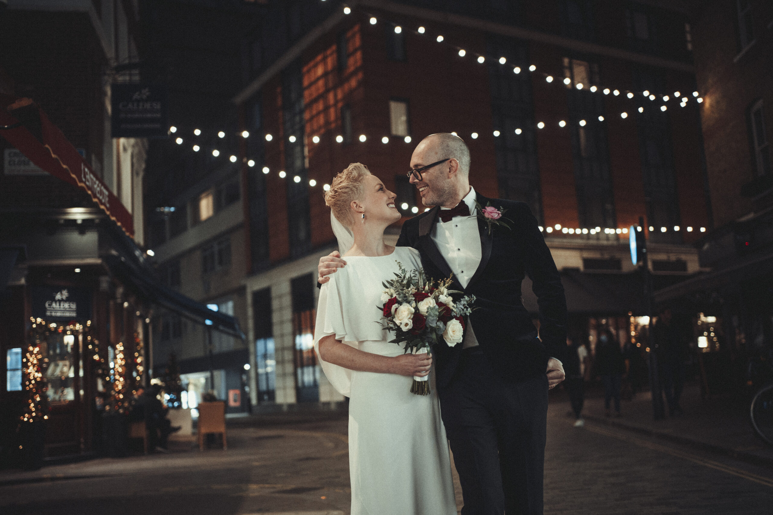 A bride and groom standing in a city street at night. (listing)